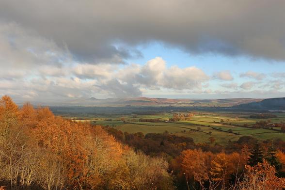 a landscape photo, the foreground is bathed in sunshine, lighting up the golden leaves of the autumn trees. In the mid ground, green fields . Beyond, moorland with patchy sunshine. The sky is full of clouds, a rapidly shrinking path of distant blue is being obscured by heavy grey cloud overhead