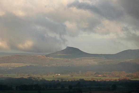 A zoomed in photo of a mostly grey landscape, very little sun is now penetrating the grey clouds. An outlying hill is silhouetted on the horizon