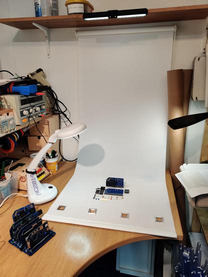 A wooden desk with electronic test equipment towards one side. On the wall at the back is a white roller blind that has been pulled down and curves on to the desk. Some electronic components are sitting on the blind with multiple light sources pointing towards it.