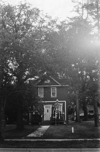 Black and white film photographs of a quiet suburban neighborhood. The first image shows a rabbit sitting alert on a neatly trimmed front lawn in front of a house. The second image shows a human skeleton decoration standing on a porch in midsummer, surrounded by typical suburban yard details.