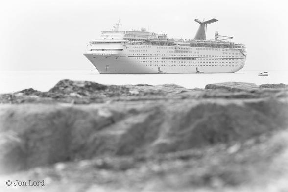 This is a hazy black and white photo of a ship at anchor in landscape format. 
The lower half of the image is filled with the very light grey rocky surface of a harbour breakwater. Above is a short stretch of dead calm ocean before the horizon is reached. Above is a white sky that may be a heavy morning haze or overcast clouds. Located an equal distance between the rocks and the horizon is a medium sized cruise ship (260m). The view is oblique with the bow and port side in view. The ship is anchored facing slightly towards the camera and filling a little more than half of the image. Other than the single 'Y' shaped funnel, rendered light grey: the rest of the ship is all white. Two rectangular hatches are open a metre or so above the waterline with two small shore boats boarding  passengers for a 'run ashore'. Above the hatches is a short row of perhaps ten round portholes. Above are six rows of rectangular windows. Next is a promenade deck with nine covered lifeboats suspended from davits. Aft of the funnel is a waterslide. On the bow and aft of the ship's bridge is the vessel's name in bold black letters 'Carnival Imagination'.
The location: Santa Catalina Island, California (2013).