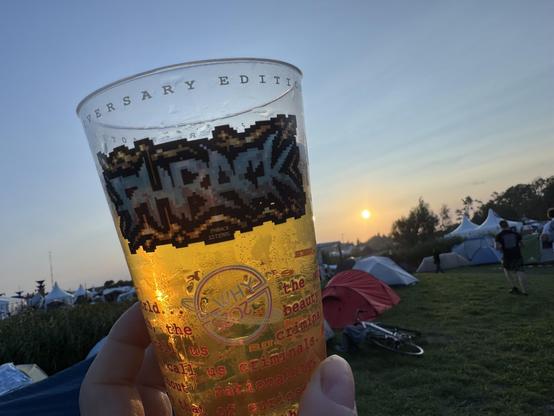 A hand holds a clear cup filled with a golden beverage, featuring a graphic label, against a sunset backdrop. In the background, tents and festival-goers are visible in a grassy area.