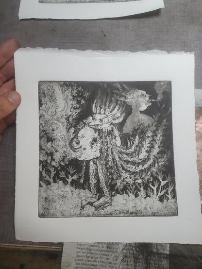 Photo, ma main tient un tirage en noir et blanc. La gravure : une grosse tête à longs cheveux tressés, bouche ouverte et yeux équarquillés, souffle des bulles / de la fumée. Elle est tenue par derrière par un personnage masqué à grand nez, dont on ne voit que la tête, le mains et les jambes. Pieds nus et jambes recouvertes d'écailles. Au sol, des silhouettes blanches de plantes, algues. Derrière la longue chevelure, une silhouette blanche, torse et tête, crache aussi quelque chose, de la fumée ?