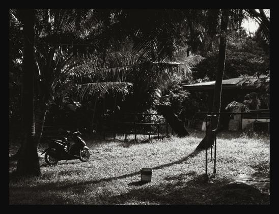 A high-contrast, black-and-white photograph captures a sunlit clearing in a tropical setting. A motorcycle is parked in the shade of dense palm trees on the left, while a lone bucket sits on the grassy ground, casting a long shadow. There's a house partially visible in the background.