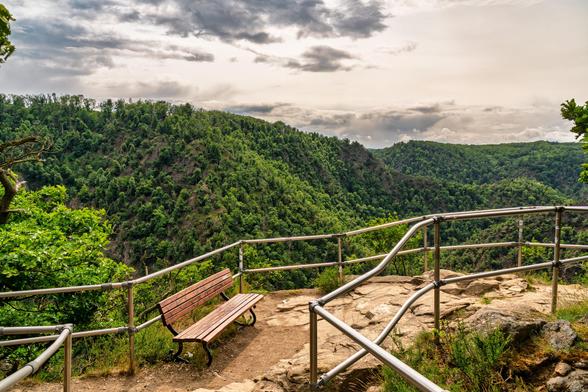 Im Vordergrund das Metallgeländer am Aussichtspunkt, das in das Bild führt. Auf der linken Seite steht eine Holzbank. Dahinter Blick auf das Bodetal und die steilen Klippen.