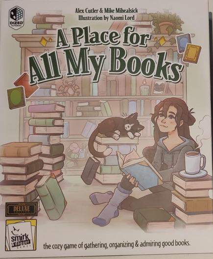 The dust cover of a board game "A place for all my books". The image shows a full bookshelf, some stacks of books, a cat sleeping on one stack, and apparently a young lady enjoying a book and a steaming  cup of coffee on another bookstack. The mood is cozy.