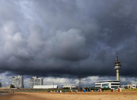 A wide-angle shot of a beach in Bremerhaven, Germany, with a dramatic, dark grey sky filled with ominous-looking storm clouds. In the foreground is a sandy beach with several colourful beach chairs. The middle ground features modern buildings, including two tall white high-rises and a white building with a prominent observation tower topped with an antenna mast. A few people are walking along the sea wall in the distance. The overall mood is slightly foreboding due to the dark, cloudy sky.