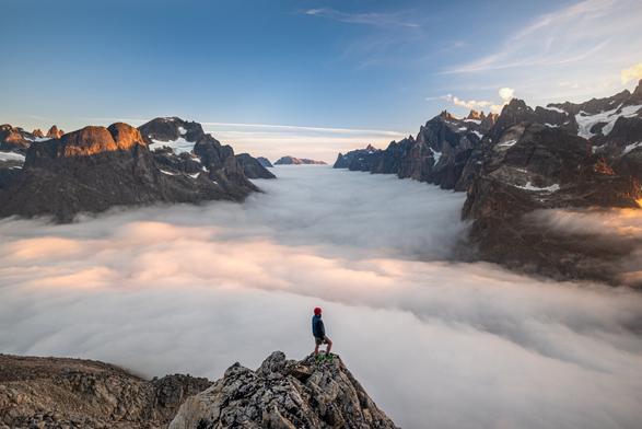 A lone hiker stands on a rugged rock outcrop at the bottom center, gazing over a vast valley filled with a thick sea of clouds; jagged mountain peaks rise on both sides, some with patches of snow, while a clear blue sky with wispy clouds spans above. The sun casts warm golden light on sunlit rocks and the hiker’s red hat and blue jacket contrast with the cool shadows and the white clouds below. The scene feels dramatic and expansive, emphasizing the hiker’s small scale against the towering, rugged landscape.