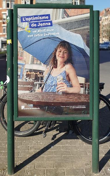 Un panneau publicitaire avec l'affiche suivante :
Une demoiselle souriante avec un parapluie à une terrasse.
Il y est inscrit :" l'optimisme de Jeanne un atout pour son futur employeur."
Affiche de l'onem.