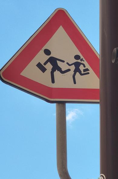 Panneau de signalisation d'enfants sortant de l'école. Triangle rouge avec 2 personnages en noir.
Fond bleu du ciel.