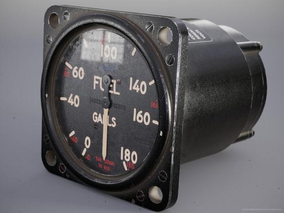 Front side view of the fuel gauge model showing the dial with divisions in gallons of fuel in the tanks and alternative values for the plane in climb. The case is dark painted anodised metal with worn edges and various fixing holes in the front there is a partially visible equipment ID label on the top of the cylindrical body.