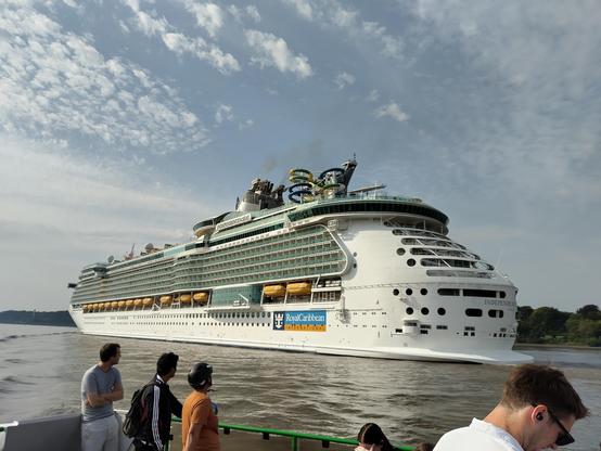 Photo showing the giant cruise ship "Independence of the Seas" sailing along the Elbe river, dwarfing the small passenger ferry from which the photo is being taken.