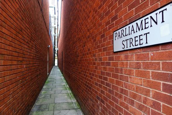 A very narrow pedestrian alleyway in Exeter, England, named Parliament Street. The path is paved with grey slabs, and the high walls on either side are made of red brick. A white street sign with black capital letters reading 'PARLIAMENT STREET' is mounted on the wall to the right. The alley is dimly lit, with a small patch of brighter light visible at the far end, suggesting an exit.