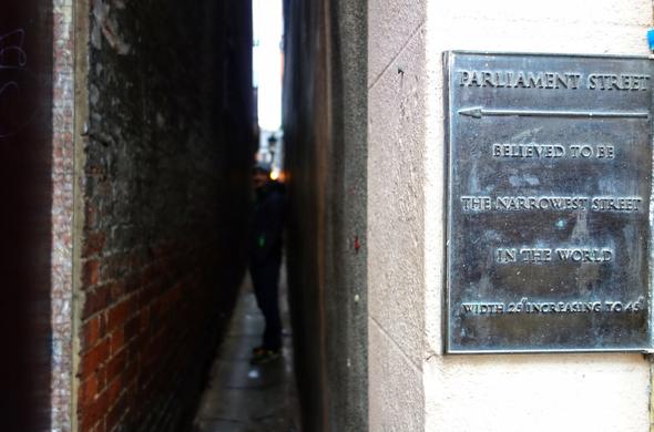 A dark, extremely narrow alleyway, Parliament Street, in Exeter, England. A person is barely visible in the middle of the passage, providing a sense of scale. A weathered bronze or brass plaque is mounted on the wall to the right, with an arrow pointing into the alley. The plaque reads 'PARLIAMENT STREET' at the top, followed by 'BELIEVED TO BE THE NARROWEST STREET IN THE WORLD' and 'WIDTH 26" INCREASING TO 45". The alley walls are made of brick and stone, and a single light source is visible at the far end.