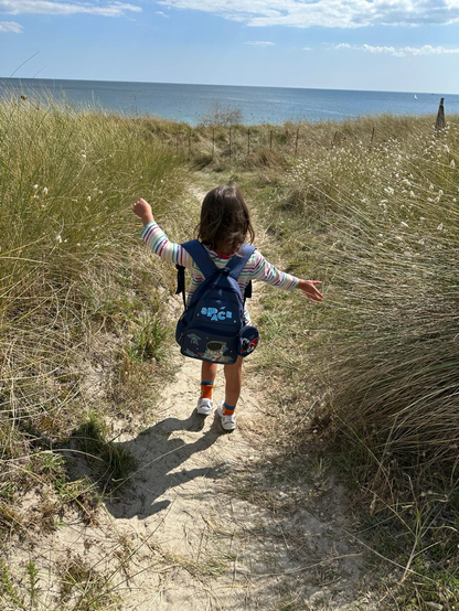 A young child with a backpack walks along a sandy path surrounded by tall grass, leading towards a beach and the ocean in the background. The child has their arms outstretched, enjoying the moment, with a bright sky above.