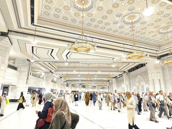 This is inside Masjid-Al-Haram (The Grand Mosque) in Saudi Arabia. It looks like a white place. Many people are walking around. There are many lights and beautiful decorative items. Everything looks grand and luxurious. The photo was taken during night time.