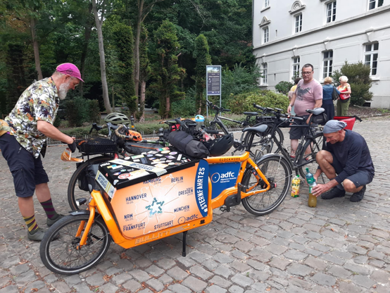 Drei Männer stehen mit Fahrrädern auf einem gepflasterten Platz vor Bäumen und einem hellen Gebäude. Im Vordergrund ist ein auffälliges, oranges Lastenrad, auf dessen Ladefläche eine Deutschlandkarte mit dem Schriftzug "#STERNFAHRT25" und "adfc" abgebildet ist. Ein Mann mit Bart und pinker Kappe steht am Lenker des Lastenrads, während ein anderer Mann daneben auf dem Boden kniet und Flaschen befüllt.