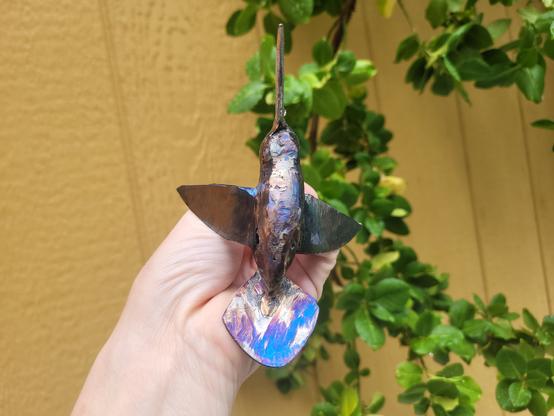 Un colibri en acier recyclé est dans une main dans un angle où l'on voit que l'acier a été coloré bleu et mauve