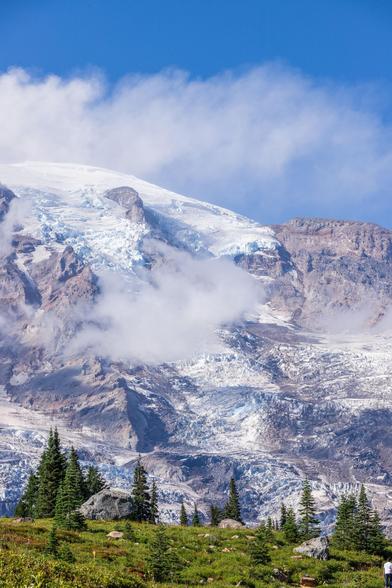 Snow-capped mountain with clouds and green trees in the foreground under a blue sky.
