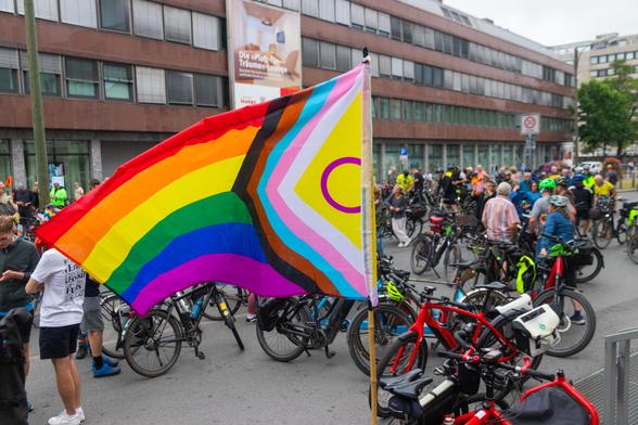 Im Vordergrund weht eine große Regenbogenfahne mit zusätzlichen Farben für Vielfalt und Inklusion. Dahinter stehen viele Menschen mit Fahrrädern auf einer Straße, einige tragen bunte Kleidung und Helme.
© Christpoher Ising