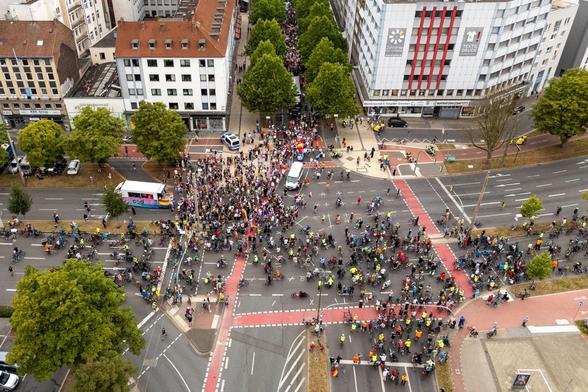 Ein Luftbild zeigt eine große Menge an Menschen und Fahrrädern, die eine Kreuzung in einer Innenstadt füllen. Inmitten der Menge fahren bunte Wagen und es sind Regenbogenfahnen zu sehen.
© Leopold Achilles