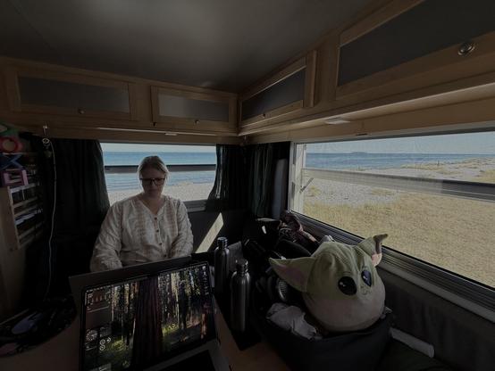 Das Bild zeigt die Innenseite eines Wohnmobils, in dem eine Person sitzt und an einem Laptop arbeitet. Das Wohnmobil hat große Fenster, durch die man einen Blick auf das Meer und den Strand hat. Die Person trägt eine weiße Bluse und hat eine Brille auf. Neben dem Laptop liegt ein großer Plüschspielzeug, das nach dem Charakter "Baby Yoda" aus der Star Wars-Serie "The Mandalorian" aussieht. Auf dem Tisch stehen zwei Thermosflaschen. Das Wohnmobil ist mit Holzverkleidungen und Regalen ausgestattet, die verschiedene Gegenstände aufbewahren. Die Szene vermittelt ein Gefühl von Mobilität und Freiheit, da das Wohnmobil an einem ruhigen Strandort geparkt ist.