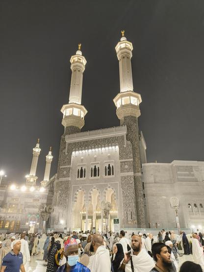 A low-angle shot of the Masjid al-Haram at night. The main focus is on a large, intricately designed gate with two tall minarets topped with golden finials, against a dark sky. The minarets and the gate are illuminated, highlighting the detailed stonework. Below, a large crowd of people, many wearing white ihram clothing, are seen walking and gathering. The atmosphere appears to be a mix of reverence and activity. The faces of some individuals in the foreground are visible.