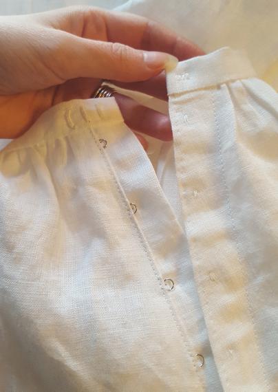 Close up photo of the back closure, a placket, on the centre back of a white linen skirt. Silver hooks and eyes have been sewn between the layers of the placket, only the active bits of the hardware visible, as well as some sewing thread affixing the hooks through all layers, quite subtle. There's a narrow waistband with three thread bars sewn at slightly different distances, one at exactly centre back, and two more one centimetre away from it, allowing for tighter and looser fit. Sini's hand holds onto the waistband. The linen is not infinitely fine, but nice looking and with some structure despite being wrinkly as heck.