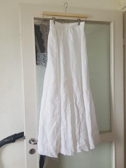 A slightly sad looking full garment image, with the skirt on a skirt hanger which is on a door. It's very narrow at the waist and significantly flares out at about mid hip, going to a sort of trumpet shape but with straight seams. It's quite wrinkled, the flat felled seams lend a tiny bit of practical mood to it, and the hem is entirely unfinished with threads coming off the weave at the raw edge. It needs to hand out like this for several days, for the fabric to let itself relax and settle to how it will want to, with gravity.