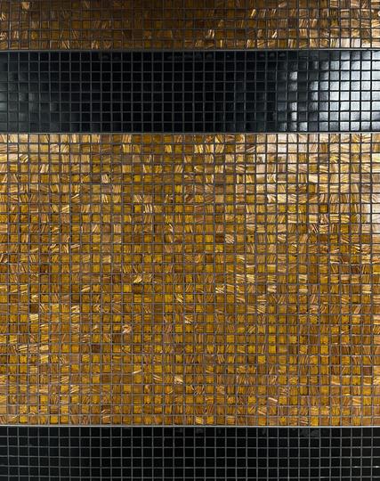 A close up shot of small square tiles. They are primarily amber, and as they reflect in the light you can see the pattern embedded within them goes in different directions. There are also two bands of black tiles, one towards the top, and one at the very bottom.