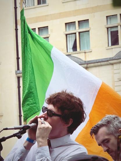 Young man with glasses playing the harmonica in front of an Irish flag