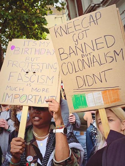 Black girl with banner saying it's her birthday but fighting fascism is more important, and blonde girl with banner "kneecap got banned but colonialism didn't"