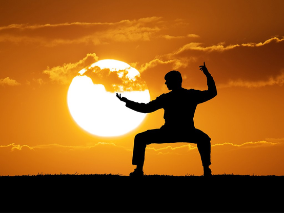 A silhouette of man practicing Tai Chi during sunset. Large sun disc going closer to the horizon, partly covered behind the clouds. Sky iwith strong blend of sunset red colors. Atmosphere is calm and relaxing.