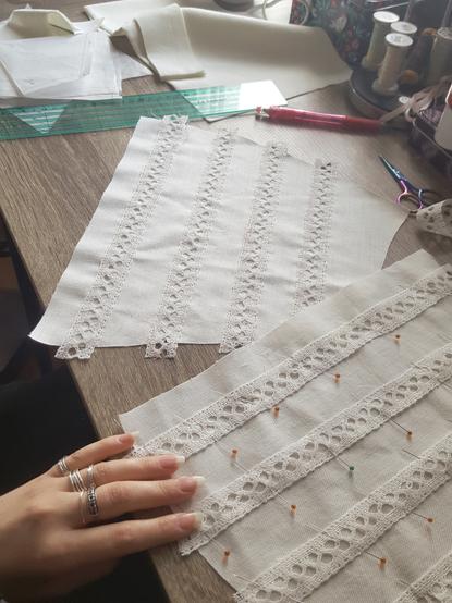 Two lower front parts of a cream blouse in progress, with ivory insertion lace pinned and hand tacked on it. There's a general disarray of other pattern pieces, scissors, pencil, ruler and such in frame, and pretty daylight from a window. The lace strips have been eyeballed into position, in vertical lines along the centre front and radiating out. Sini's hand for scale and colour matching, the fabric looking a bit more white than it actually is.
