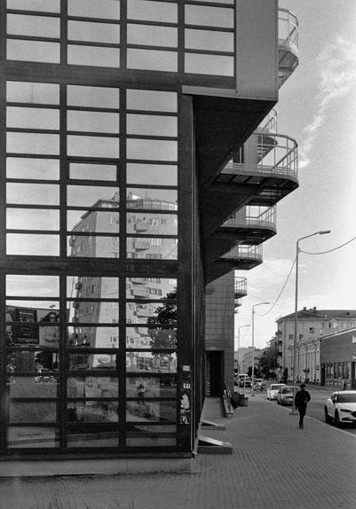 A black-and-white photograph of a city street with modern architecture. On the left side of the frame, there is a building with large square windows that reflect the building opposite. The building is flanked by open staircases with circular balconies. To the right of the building, there is a sidewalk with a silhouette of a person walking on it. Cars are parked along the street, and other buildings can be seen in the distance.