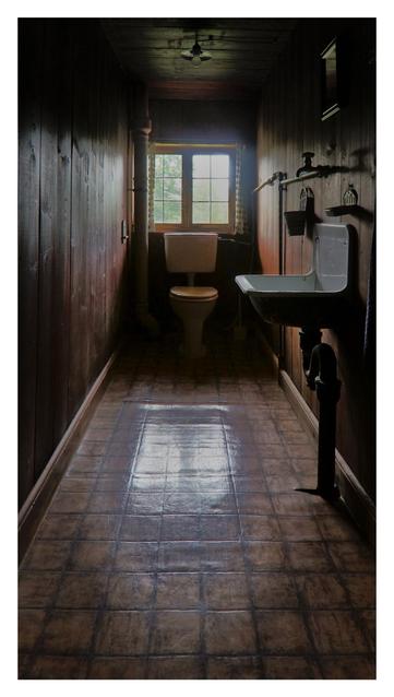 This image depicts a long narrow, dimly lit bathroom with a rustic, vintage feel. The room has dark wooden walls and a tiled floor with a glossy finish, reflecting some light. On the right side, there is a white sink with a black pipe and a small shelf above it. Further down, a white toilet with a wooden seat is positioned against the wall. A small window with a grid pattern at the end of the room allows natural light to stream in, illuminating the space and casting a soft glow on the floor. On the left wall, there’s a vertical pipe, and on the right wall, a mirror and some hanging items, possibly towels or decorations, are visible. The overall atmosphere is moody and old-fashioned, with the light creating a contrast between the bright window area and the shadowy corners.