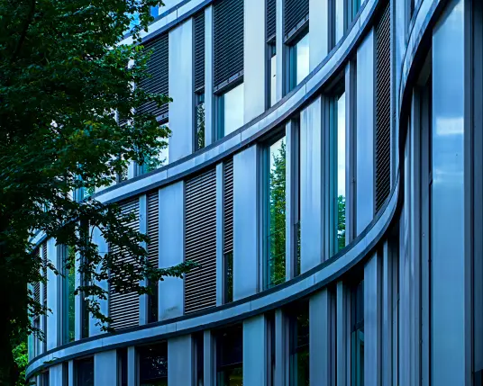 Drei Stockwerke voller Fenster schwingen sich in einer S-Form von rechts nach links durch das Bild. Die Fenster wechseln sich in jeder Reihe mit glatten Fassadenplatten ab. Die S-Form ergibt sich aus drei schmalen Streifen, die zwischen den Stockwerken entlanglaufen. 
Von links ragt ein dichter, grüner Baum in das Bild. Das ganze Gebäude macht einen kühlen, sehr modernen Eindruck. 
. 
Three floors full of windows swing in an S-shape from right to left across the photo. The windows alternate in each row with smooth front panels. The S-shape is made up of three narrow strips that run between the floors. A dense, green tree protrudes into the photo from the left. The whole building gives a cool, very modern impression.
.