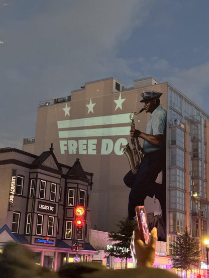 A nighttime street scene in Washington D.C. at the intersection of 14th Street NW and U Street NW. A large projection displays "FREE DC" with three stars above it (mimicking the D.C. flag design) on the side of a brick building. To the right is a prominent mural of Buck Hill, known as "The Wailin' Mailman," depicted as a Black man in a postal worker's cap playing a saxophone. The scene shows typical D.C. urban architecture with historic rowhouses on the left and modern apartment buildings on the right.