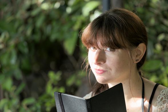 Portrait rapproché d’une brune. Elle tiens un carnet noir qui dépasse du cadre et regarde la caméra avec une étincelle dans les yeux, comme si elle venait d’avoir une idée. Le fond est une haie verte