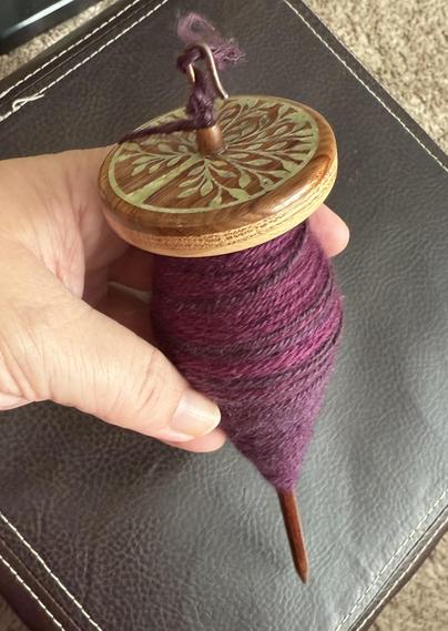 Drop spindle with a full cop of purple tonal yarn. The spindle whorl has a beautiful inlaid tree, light green against the medium brown grain of the wood