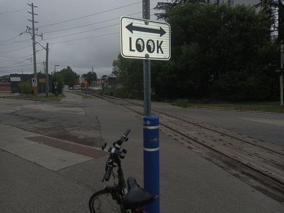 Looking down the Spur Line Trail, where a sign cautions trail users to "LOOK". The O's have bloodshot eyeballs painted in, looking down at the startled bollard and Bob's bike. The Spur Line tracks recede into the distance to the right of the trail, which crosses Union Street here.