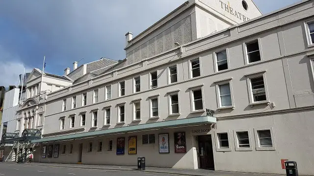 The image depicts the exterior of a theater building. It features a beige facade with multiple windows, a green awning over the entrance labeled "STAGE DOOR," and various posters displayed on the walls. The sky is partly cloudy, and the surrounding area looks urban.

Image Credits: Wikimedia / Stinglehammer / CC BY-SA 4.0