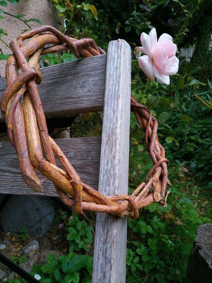 Über einer Lehne eines alten Klappstuhls hängt ein geflochtener Holzring aus Weide. Dahinter eine rosa Rosenblüte.