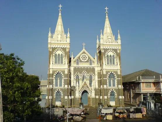 The image features a prominent church with distinctive gothic architectural elements, including two tall spires and elaborate stonework. The façade displays intricate designs, and the setting appears to include nearby buildings and structures, possibly indicating a community or market area. The sky is clear and blue, suggesting a bright day.

Image Credits: Wikimedia / Rakesh Krishna Kumar / CC BY-SA 2.0