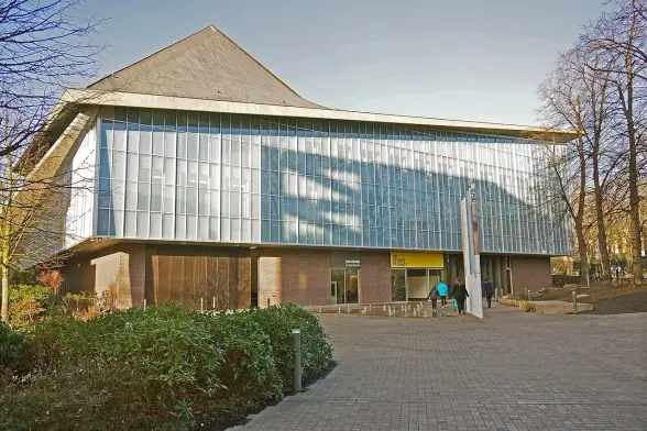 The image appears to showcase a modern building with a distinctive architectural design. The structure features a glass façade that reflects its surroundings, with a sloped roof. In front of the building, there seems to be a paved pathway and some greenery, possibly a landscaped area. People can be seen walking towards the entrance, which is marked by a yellow section. The overall setting suggests it could be an educational or community facility.

Image Credits: Wikimedia / Anthony O'Neil / CC BY-SA 2.0
