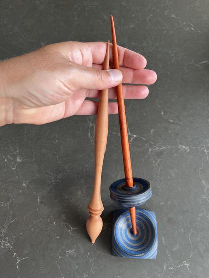 A Russian style spindle made of pear wood and a Tibetan style spindle made of reddish padouk wood with a blue whirl and a blue spinning bowl. They are support spindles and held loosely in a left hand.