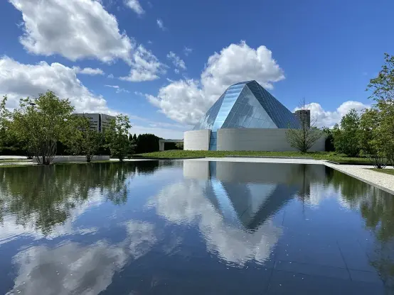 The image features a geometric glass structure, likely a museum or gallery, reflecting in a body of water. The setting includes green landscaping and a clear blue sky with scattered clouds. The combination of the reflective pool and the architecture creates a visually appealing scene.

Image Credits: Wikimedia / Canmenwalker / CC BY 4.0