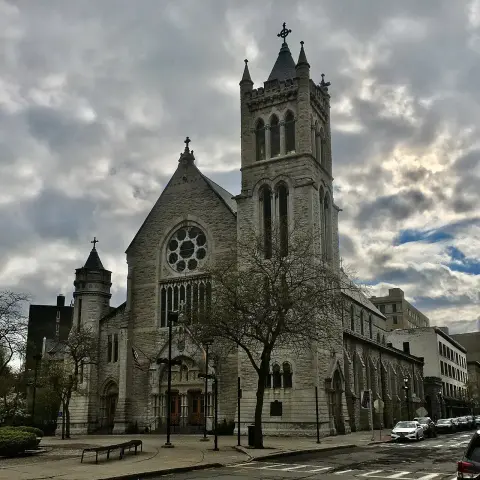 The image shows a large church building with distinctive architectural features, including a tall bell tower, large stained glass windows, and a stone facade. The sky appears cloudy, adding to the atmosphere of the scene. Trees and parked cars are also visible in the foreground and surrounding area.

Image Credits: Wikimedia / Andre Carrotflower / CC BY-SA 4.0