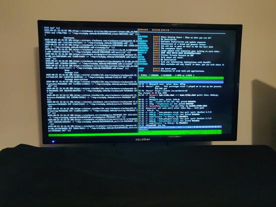 My HDTV connected as a monitor to my desktop PC. On the screen is Slackware Linux in console mode with Tmux running a two panes, side by side. Left pane is running slackpkg+ updates and upgrades. Right pane is SSH'd to tilde.institute and running a Tmux session there, split into two panes, top and bottom. Bottom pane is showing Irssi in an IRC channel. Top pane is SSH'd to SDF and running another Tmux session there, and focused on the Tmux window where the list of BBOARD boards is shown.