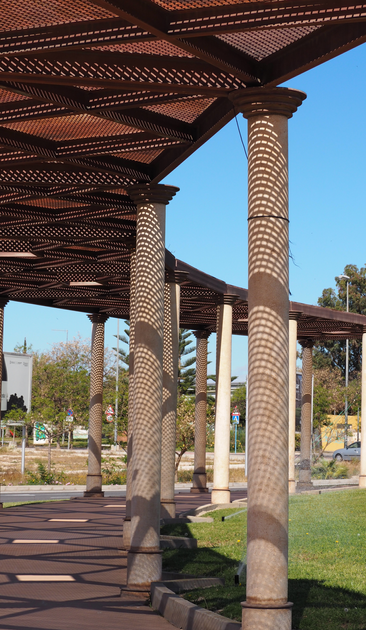 Eine bogenförmige Pergola mit runden Säulen und einem Dach aus T-Trägern und Gittern erzeugt interessante Schattenspiele am Boden und den Säulen. Im Hintergrund Fahrbahnen, Bäume, Plakate und blauer Himmel.